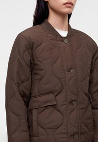 Personne portant une veste matelassée marron avec fermeture à boutons, col côtelé, poches à rabat et motif cousu ondulé.