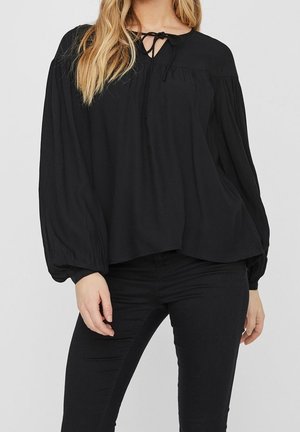 Vrouw die een losse zwarte blouse draagt met lange pofmouwen en een strikdetail bij de halslijn, gecombineerd met nauwsluitende zwarte broek.