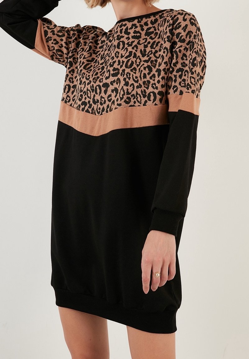Femme portant une robe noire à manches longues avec un imprimé léopard et une bande horizontale marron traversant la poitrine et le haut des bras.