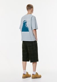 Person står med hænderne i lommerne iført en lyseblå t-shirt med grafik, mørke oversized shorts, hvide sokker og brune vandrestøvler.