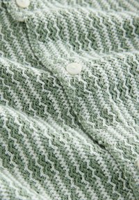 Svijetlozelena pletenina s teksturiranim cik-cak uzorkom, s malim bijelim gumbima duž ruba. Mekan i visoko rastezljiv materijal.