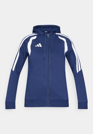 Granatowa bluza z kapturem zapinana na zamek, z troczkiem przy kapturze, białymi paskami po bokach i logo Adidas na górnej, lewej stronie klatki piersiowej. Miękka struktura tkaniny.