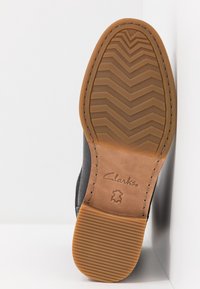 Brązowa gumowa podeszwa z zygzakowym bieżnikiem i stebnowanym brzegiem. Ciemny materiał górny częściowo widoczny. Zawiera logo Clarks na pięcie.