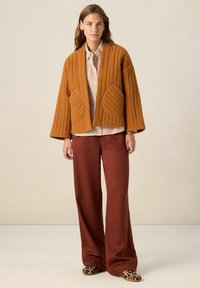 Veste matelassée marron avec de grandes poches à côtes, associée à une chemise à rayures à boutons et un pantalon marron à jambes larges, portée avec des chaussures à motifs.