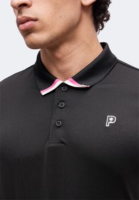 Jeune homme portant un polo noir à trois boutons, un col avec des rayures roses et blanches, et un petit logo "P" avec un palmier.