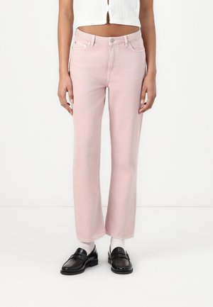 Femme portant un jean droit rose clair associé à des mocassins noirs et des chaussettes blanches, debout devant un fond blanc uni.