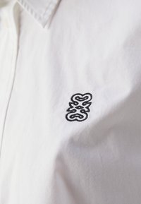 Camisa de algodón blanca que presenta un destacado logo bordado en negro en el lado izquierdo del pecho, con un cuello clásico y una textura suave.