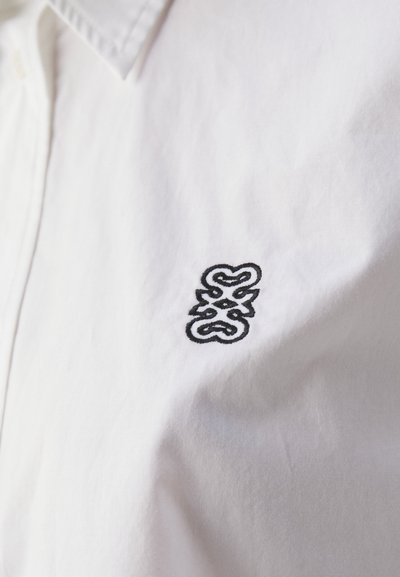 White cotton shirt featuring a prominent black embroidered logo on the left chest, with a classic collar and smooth texture.