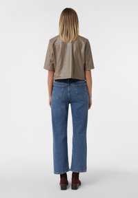 Gekürztes taupefarbenes Lederoberteil mit kurzen Ärmeln, kombiniert mit hochgeschnittenen blauen Jeans und braunen hohen Stiefeln. Einfarbiger Hintergrund.
