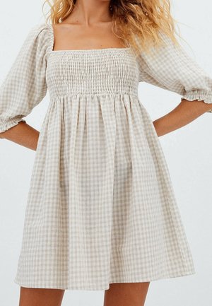 Femme portant une robe à carreaux beige et blanche avec des manches trois-quarts bouffantes et un décolleté carré froncé.