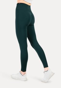 Leggings verde oscuro con una cinturilla alta, hechos de un material elástico. Un par de zapatillas deportivas blancas con detalles texturizados visibles.