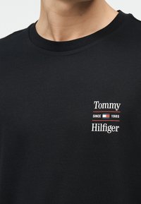 Κοντινό πλάνο ενός ατόμου που φοράει μαύρο T-shirt με το κείμενο "Tommy Hilfiger Since 1985" και ένα μικρό κόκκινο, λευκό και μπλε λογότυπο στο στήθος.