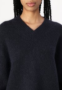 Pull en tricot noir texturé avec un col en V, porté par une personne aux cheveux tressés visibles sur un fond blanc.