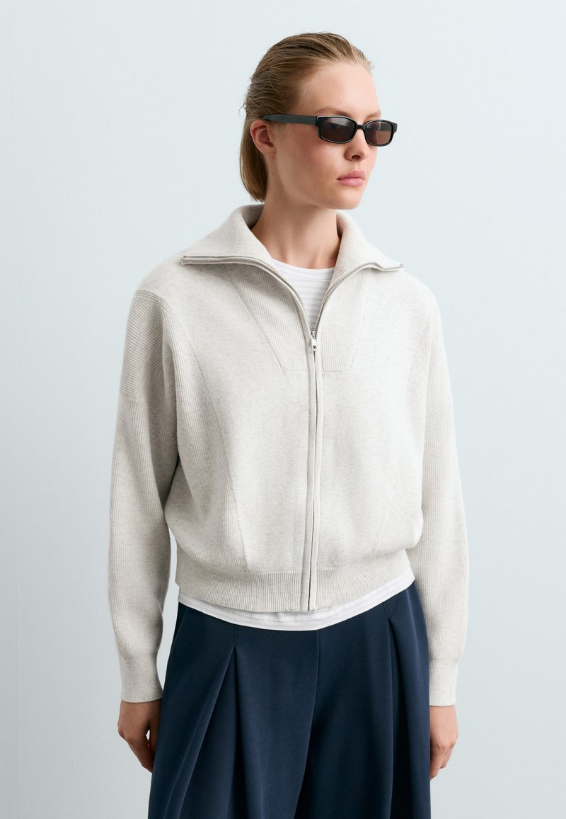 Femme aux cheveux blonds attachés, portant des lunettes de soleil noires, un pull beige clair zippé, un t-shirt blanc en dessous et un pantalon plissé bleu foncé.