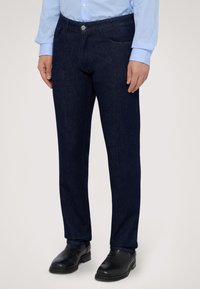 Pantalones de mezclilla oscura con corte recto, que cuentan con un clásico diseño de cinco bolsillos y un cierre de botón plateado, combinados con zapatos negros.