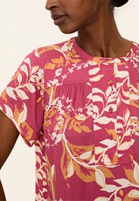 Blusa con motivo floreale nei colori rosa, arancione e bianco. Presenta un collo rotondo, maniche corte e dettagli con bottoni. Tessuto testurizzato con pieghe.