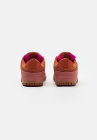 Un paio di sneakers arancioni e rosa viste da dietro, con interno rosa e suole in gomma marrone su uno sfondo bianco.