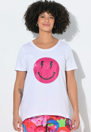 Mujer con cabello rizado que lleva una camiseta blanca con una cara sonriente rosa y ojos en forma de rayo, combinada con pantalones con estampado colorido.