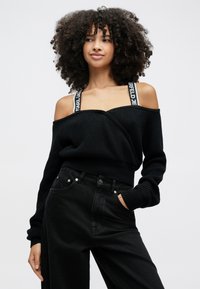 Kvinna med lockigt hår som bär en svart off-shoulder tröja med logoband och höga svarta byxor, poserande med handen i fickan.