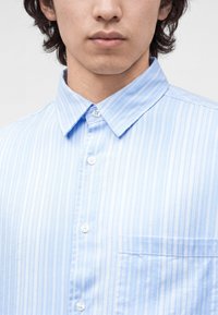 Jeune personne aux cheveux mi-longs foncés porte une chemise à rayures verticales bleu clair et blanche avec une poche poitrine.