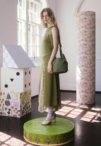 Olivově zelené šaty s květinovými akcenty, doplněné o zelenou kabelku a žluté sandály na podpatku. Modelka stojí na kruhové zelené podložce.