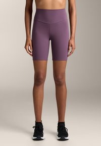 Magas derékú lila kerékpáros nadrág, rugalmas anyagból készült, sima textúrájú és térdhosszúságú, fekete sportcipőkkel párosítva.