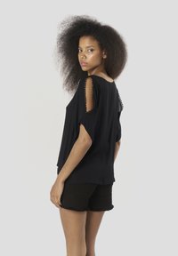 Top negro ligero con mangas cortas abiertas, con adorno decorativo a lo largo de las costuras de los hombros y un corte holgado. Combinado con pantalones cortos negros.