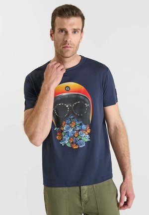 Mann trägt ein navyblaues T-Shirt mit einem Grafikdesign von einem Helm, Sonnenbrillen und Blumen mit Schmetterlingen, kombiniert mit grünen Hosen.