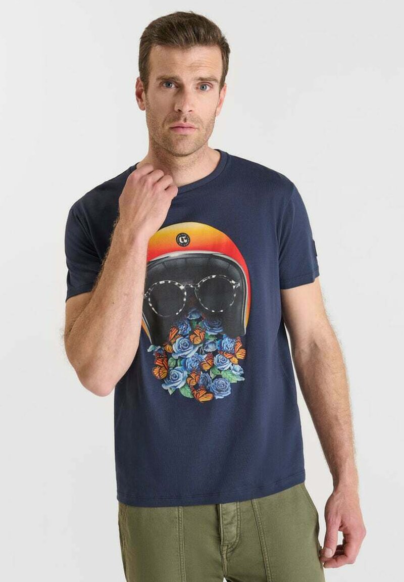 Hombre con una camiseta azul marino que presenta un gráfico de un casco, gafas de sol y flores con mariposas, combinada con pantalones verdes.