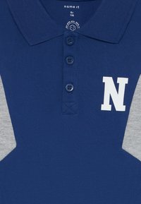 Marineblå polo-skjorte med to grå sidepaneler, tre-knapp placket og en hvit "N" trykt på forsiden. Laget av teksturert materiale.