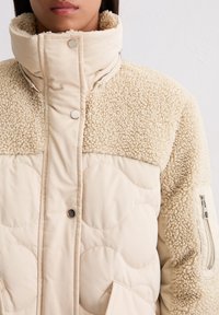 Frau trägt beige gesteppte Winterjacke mit Sherpa-Futter, hohem Kragen und Druckknöpfen, sichtbare Reißverschlusstasche am Ärmel.