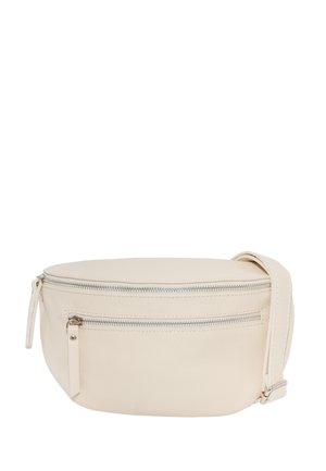 Frau mit langen braunen Haaren, die ein schwarzes Langarmoberteil und dunkle Jeans trägt und eine beige Umhängetasche quer über der Brust hält.