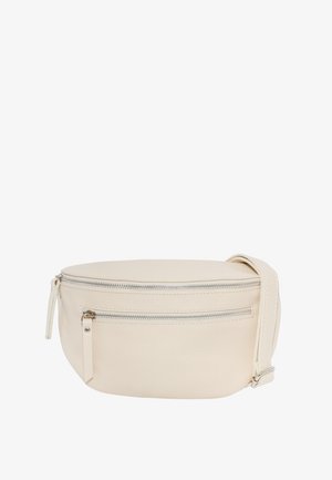 Frau mit langen braunen Haaren, die ein schwarzes Langarmoberteil und dunkle Jeans trägt und eine beige Umhängetasche quer über der Brust hält.