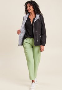 Schwarze Zip-Jacke mit einem gemusterten Innenfutter, kombiniert mit hellgrünen Cargo-Hosen mit Seitentaschen und weißen Sneakers.