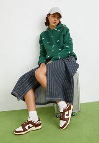 Green hoodie with white branded text, striped denim midi skirt, and brown Nike sneakers. White socks visible. Light grey cap. Seated on a metal box.