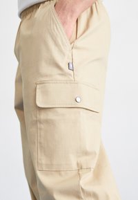 Beige Cargo-Hosen mit einem elastischen Bund, seitlichen Taschen, Druckknopfverschluss und einem kleinen Logotag. Glatte Stofftextur mit einem lockeren Schnitt.