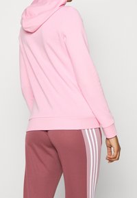Jasnoróżowa bluza z kapturem ze sznurkiem, ze ściągaczami na rękawach i u dołu, w zestawie z różowymi spodniami dresowymi z białymi paskami po bokach. Miękka tekstura materiału.