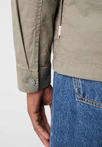 Main droite d'une personne portant une chemise beige à manches longues avec poignets boutonnés et un jean en denim bleu avec une couture visible sur la poche arrière.