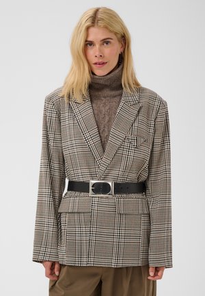 Femme aux longs cheveux blonds porte un col roulé marron, un blazer à carreaux ceinturé et un pantalon marron, debout devant un fond clair et uni.