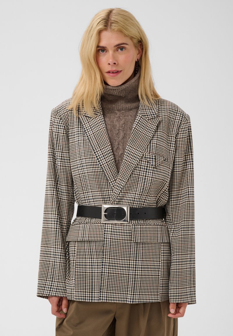 Femme aux longs cheveux blonds porte un col roulé marron, un blazer à carreaux ceinturé et un pantalon marron, debout devant un fond clair et uni.