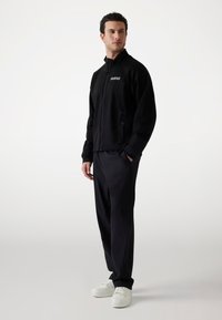 Svart zip-up jacka med hög krage, två sidofickor och "GUESS"-logotyp. Kombinerad med svarta byxor och vita sneakers med en strömlinjeformad design.
