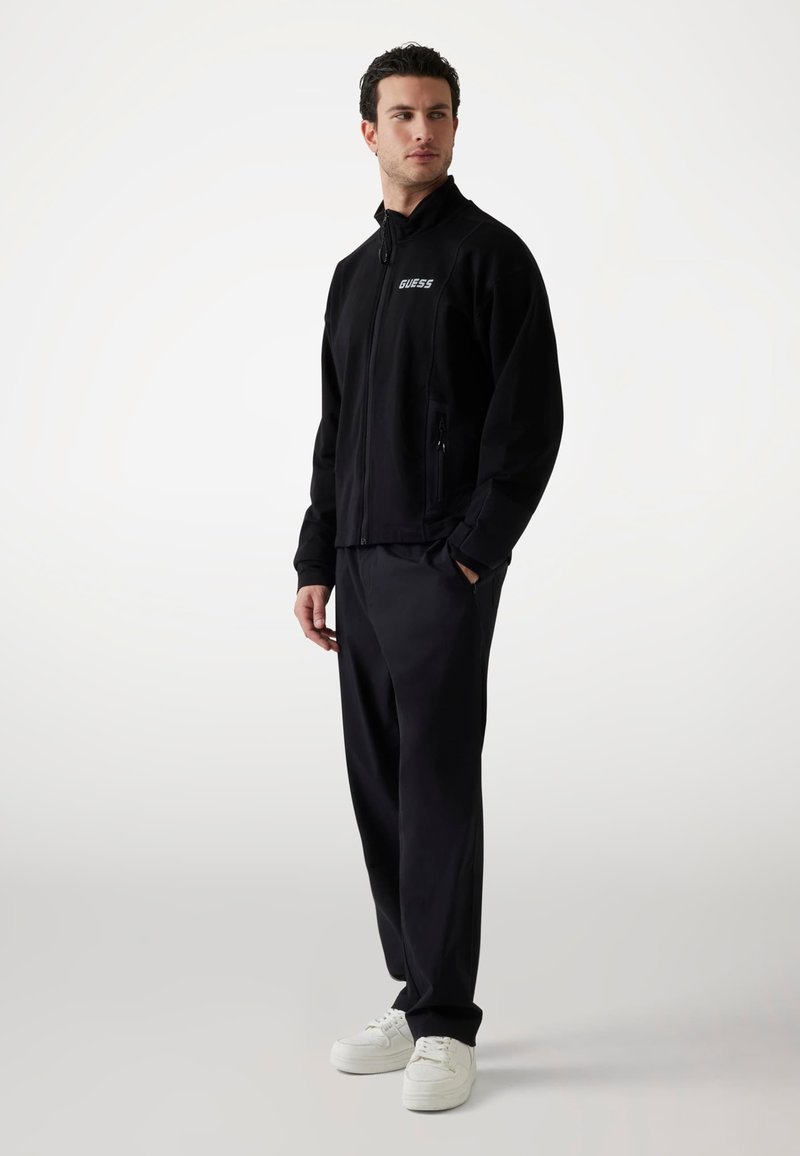 Zwarte zip-up jas met hoge kraag, twee zijzakken en het "GUESS" logo. Gecombineerd met zwarte broek en witte sneakers, met een gestroomlijnd ontwerp.