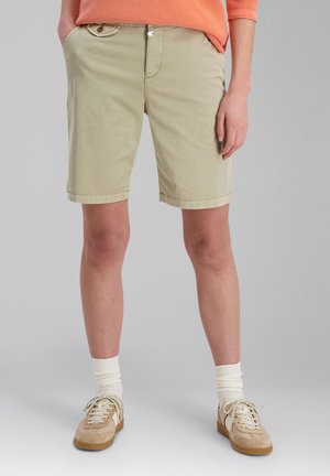 Person wearing beige knee-length shorts, white socks, beige sneakers, and an orange long-sleeve top with hands in pockets.