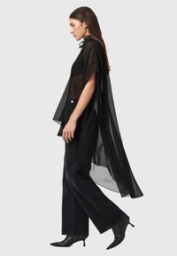 Kobieta idąca w przezroczystej czarnej, zwiewnej bluzce, szerokich czarnych spodniach i czarnych szpilkach z ostrym czubkiem, na tle gładkiego jasnego tła.