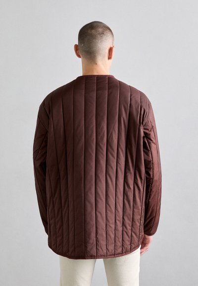 Brown quilted jacket with vertical stitching, long sleeves, and a round neck. Smooth texture with a relaxed fit, paired with light-colored pants.