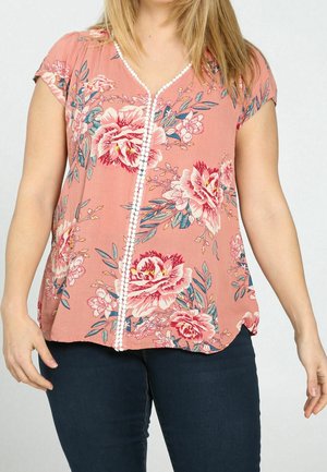 Kobieta ubrana w różową bluzkę z krótkim rękawem, ozdobioną dużymi kwiatowymi wzorami i białym obszyciem, w połączeniu z granatowymi dżinsami.