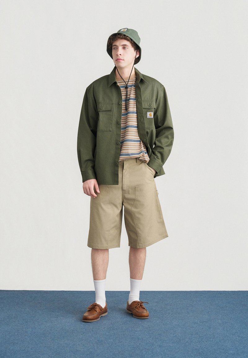 Chemise verte à boutons, chemise à manches courtes rayée en dessous, short beige, chaussures bateau marron, chaussettes blanches, chapeau seau vert. Fond minimaliste.