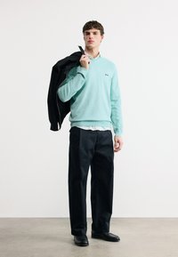 Jeune homme portant un pull bleu clair et un pantalon noir, tenant une veste noire sur son épaule, debout sur un fond uni.