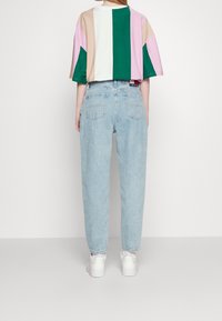 Person som bär ljusblå hög midja jeans, vita sneakers och en lös, kortärmad skjorta med vertikala färgblock i grönt, rosa, beige och vit.