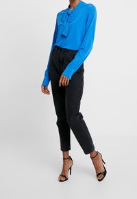 Blå långärmad blus med knytning vid halsen, kombinerad med högmidjade svarta jeans och svarta högklackade sandaler. Slät tyg.